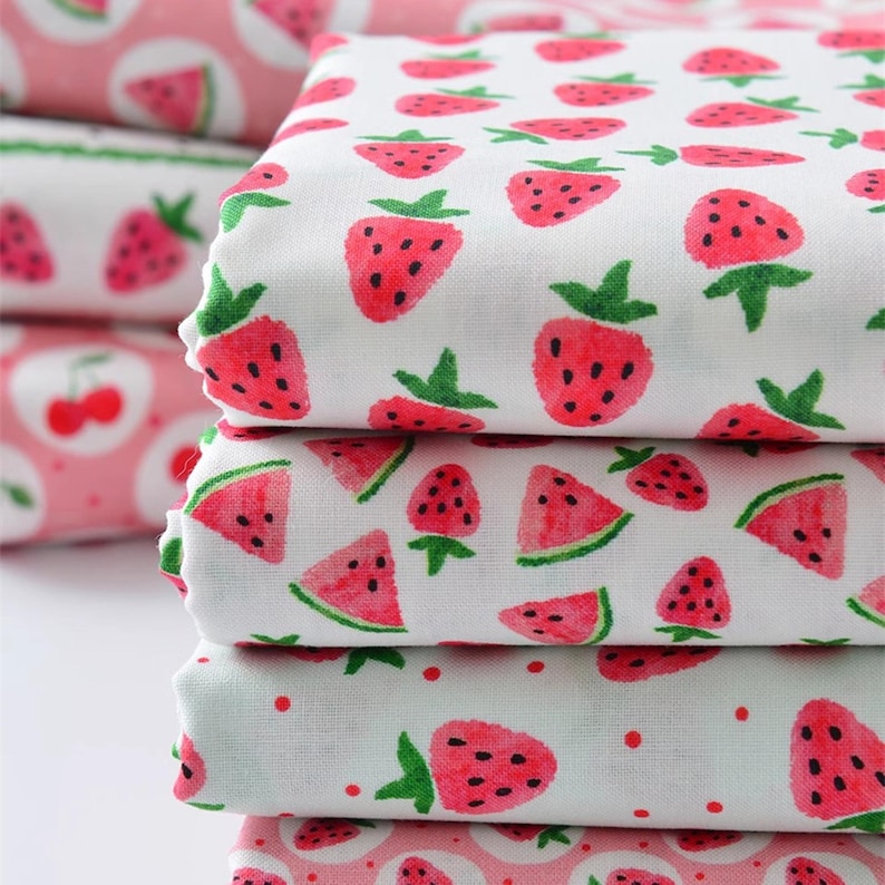 Puede incluir: Una pila de tela blanca doblada con un patr&oacute;n repetido de fresas rojas y rodajas de sand&iacute;a. Las fresas tienen tallos verdes y semillas negras. Tambi&eacute;n se ve tela adicional con un patr&oacute;n de cerezas rosas.