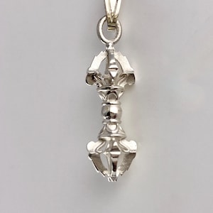 May include: A silver pendant with an intricate, symmetrical design. The pendant has a loop for a chain and a decorative, ornate shape. The design is likely symbolic, possibly religious or spiritual in nature, and is made of a shiny metal.