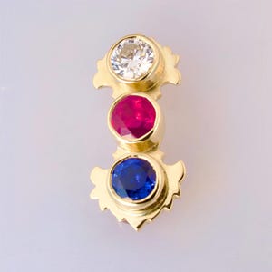 May include: A gold pendant with three gemstones: a large round diamond, a smaller round ruby, and a smaller round sapphire.