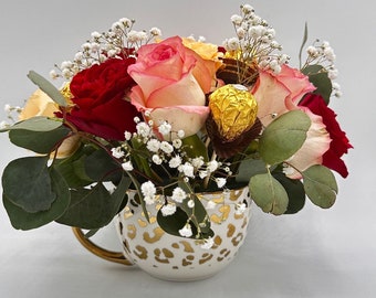 May include: A bouquet of pink, red, and white roses with baby's breath arranged in a white ceramic teacup with a gold leopard print design.