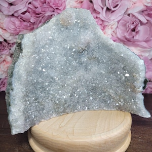 Può includere: Una grande geode di cristallo grigio chiaro con scintillanti formazioni cristalline. La geode è esposta su un piedistallo di legno chiaro, sullo sfondo di fiori di carta rosa e viola. È un oggetto decorativo naturale.
