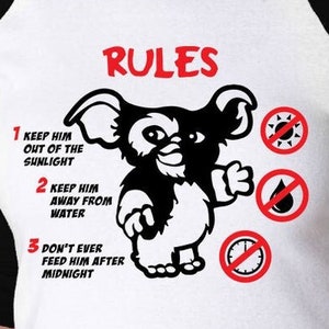 May include: A white and black baseball t-shirt with a black and white illustration of a furry creature with the text "RULES" above the creature and a list of rules below the creature. The rules are: 1. Keep him out of the sunlight. 2. Keep him away from water. 3. Don't ever feed him after midnight.