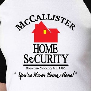 Puede incluir: Camiseta de béisbol blanca y negra con un gráfico de casa roja y el texto "MCCALLISTER HOME SECURITY FOUNDED CHICAGO, ILL. 1990 "You're Never Home Alone!"