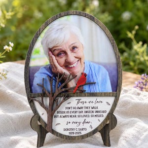 May include: An oval wooden memorial plaque featuring a photograph of an older person with white hair. A dark brown wooden tree cutout with a small red cardinal bird is attached to the front. The text reads: Those we love, DON'T GO AWAY THEY WALK BESIDE US EVERY DAY. UNSEEN, UNHEARD BUT ALWAYS NEAR. SO LOVED, SO MISSED. so very dear. DOROTHY C. SARTO 1928-2025.