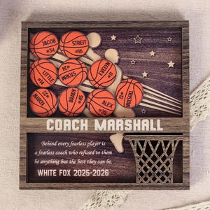 May include: Wooden basketball-themed plaque with a dark wood frame. Features basketballs with player names and numbers, a basketball net, and the text "COACH MARSHALL". Includes the text "WHITE FOX 2025-2026".