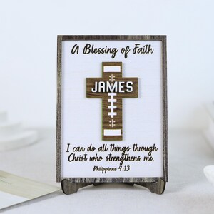 May include: A wooden framed sign with the text "A Blessing of Faith" and "I can do all things through Christ who strengthens me. Philippians 4:13." A wooden cross with the name "JAMES" is in the center.
