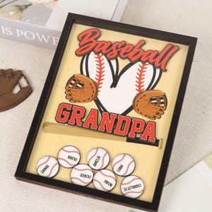 Peut inclure: Un souvenir sur le th&egrave;me du baseball encadr&eacute; avec le mot "Baseball" en script rouge au-dessus d'un c&oelig;ur en forme de balle de baseball. En dessous, le mot "Grandpa" est en lettres rouges. Comprend des balles de baseball avec des noms et une batte en bois.