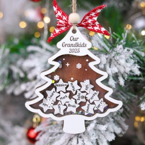 Könnte beinhalten: Ein weihnachtsbaumförmiger Ornament mit dem Text "Our Grandkids 2025". Das Ornament ist weiß mit braunem Innenraum und sternförmigen Aussparungen mit Namen. Es ist mit einer rot-weißen Schleife verziert und hängt an einer Schnur.