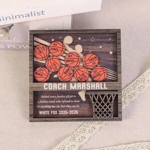May include: Wooden basketball-themed plaque with a dark brown frame. The plaque features orange basketballs with player names and numbers, a basketball net, and the text "COACH MARSHALL" and "WHITE FOX 2025-2026".