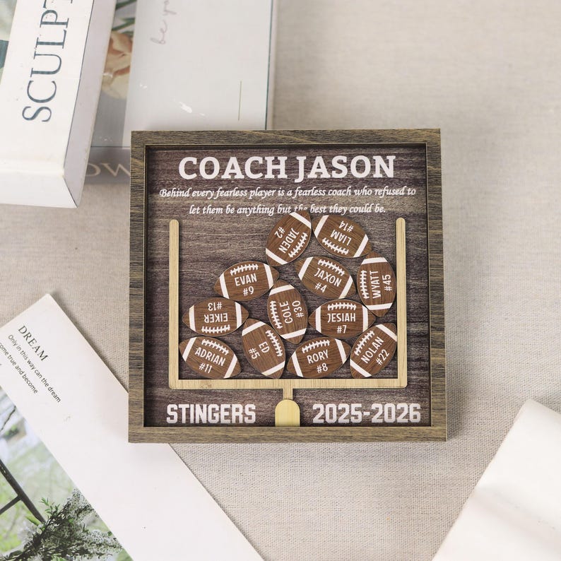 Może przedstawiać: Kwadratowa drewniana ramka typu shadow box z tłem o wzorze ciemnego drewna i białą grafiką słupk&oacute;w bramki. Zawiera napis COACH JASON, STINGERS, 2025-2026 oraz cytat. Dołączono jedenaście małych drewnianych wycięć w kształcie piłek do futbolu, każde z imieniem i numerem.