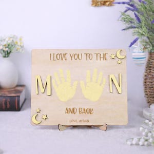 May include: Wooden plaque with the phrase "I LOVE YOU TO THE MOON AND BACK" and handprints. The plaque includes the name "AYDAN" at the bottom. Decorative stars and a crescent moon are also included.