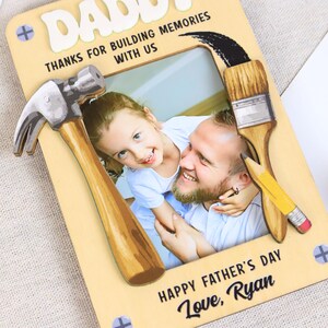 May include: A Father's Day photo frame with the word "DADDY" at the top. The frame has a hammer, paintbrush, and pencil design. The text reads "THANKS FOR BUILDING MEMORIES WITH US" and "HAPPY FATHER'S DAY Love, Ryan."