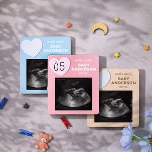 May include: Three baby announcement frames in blue, pink, and beige, each with a sonogram image. The frames have heart-shaped accents and text that reads "weeks until BABY ANDERSON arrives". A small teddy bear and decorative stars are scattered around.