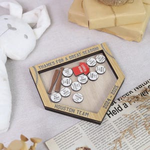 K&ouml;nnte beinhalten: Eine Baseball-Schattenbox aus Holz mit dem Text "THANKS FOR A GREAT SEASON!" und "HOUSTON TEAM". Sie enth&auml;lt Miniatur-Baseb&auml;lle mit Spielernamen und -nummern, zwei Holzschl&auml;ger und ein rotes Etikett mit der Aufschrift "COACH BROWN". Das Jahr 2024-2025 wird ebenfalls angezeigt.