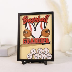 Peut inclure: Un coffret souvenir sur le th&egrave;me du baseball avec un cadre noir et les mots "Baseball Grandpa" en rouge et blanc. La bo&icirc;te pr&eacute;sente des balles de baseball, des gants et des balles personnalis&eacute;es.