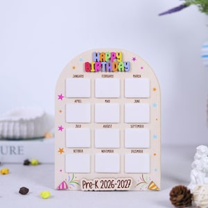 May include: Wooden birthday calendar with the words "HAPPY BIRTHDAY" in colorful letters. The calendar has 12 blank rectangular spaces, one for each month, and is decorated with stars and the text "Pre-K 2026-2027".