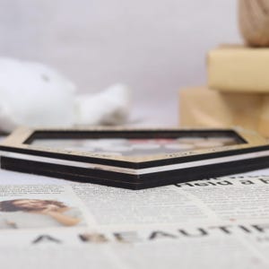 K&ouml;nnte beinhalten: Ein Holzbilderrahmen mit einem schwarz-wei&szlig;en Rand. Der Rahmen liegt auf einer Zeitung. Der Rahmen hat eine transparente Glas- oder Kunststoffabdeckung. Der Rahmen ist hellbraun.