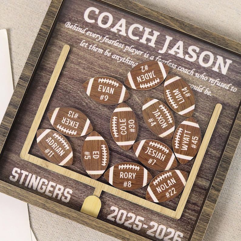 Może przedstawiać: Drewniana ramka typu shadow box z motywem boiska do futbolu amerykańskiego, z napisem COACH JASON oraz STINGERS 2025-2026. Wewnątrz znajduje się jedenaście małych, drewnianych wycięć w kształcie piłek, spersonalizowanych imieniem i numerem zawodnika.