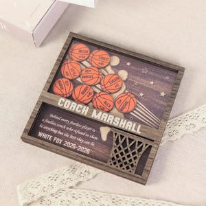 May include: Wooden basketball-themed plaque with laser-cut details. Features orange basketballs with player names and numbers, a basketball hoop, and text that reads "COACH MARSHALL" and "WHITE FOX 2025-2026". The background has a star and stripe design.