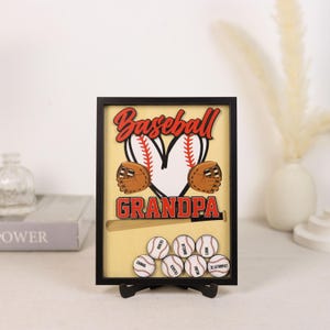 Peut inclure: Une &oelig;uvre d'art encadr&eacute;e sur le th&egrave;me du baseball. Le mot "Baseball" est en &eacute;criture rouge au-dessus d'un motif de baseball en forme de c&oelig;ur. En dessous, le mot "Grandpa" est en lettres rouges. Comprend des balles de baseball avec des noms.