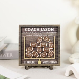 Może przedstawiać: Kwadratowa drewniana ramka z ciemnym usłojeniem, zawierająca napis COACH JASON, STINGERS oraz 2025-2026. Złota bramka podtrzymuje kilka małych, drewnianych wycięć w kształcie piłek do futbolu amerykańskiego z wygrawerowanymi imionami i numerami.