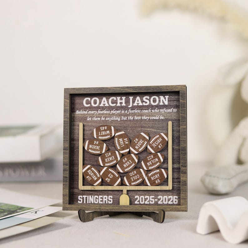 Może przedstawiać: Kwadratowa drewniana ramka z ciemnym drewnianym tłem i wyciętą z jasnego drewna bramką. Zawiera napis COACH JASON, STINGERS, 2025-2026 oraz cytat. Dołączono jedenaście małych drewnianych wycięć w kształcie piłek do futbolu z imionami i numerami.
