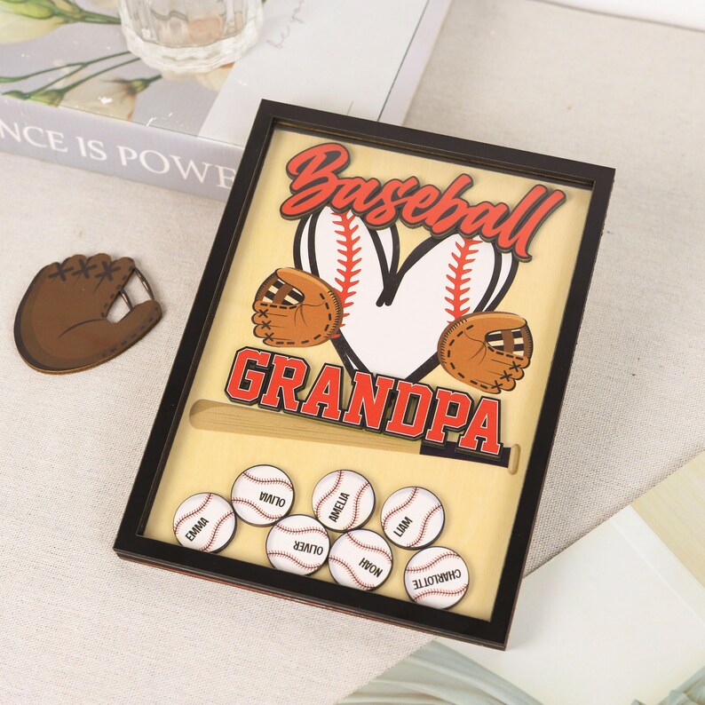 Peut inclure: Un souvenir sur le th&egrave;me du baseball encadr&eacute; avec le mot "Baseball" en script rouge au-dessus d'un c&oelig;ur en forme de balle de baseball. En dessous, le mot "Grandpa" est en lettres rouges. Comprend des balles de baseball avec des noms et une batte en bois.