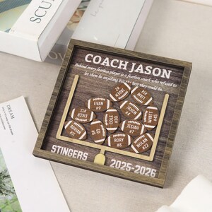 Może przedstawiać: Kwadratowa drewniana ramka typu shadow box z motywem boiska do futbolu amerykańskiego i bramką. Zawiera napis COACH JASON, STINGERS, 2025-2026 oraz cytat. Wewnątrz znajduje się jedenaście małych, drewnianych wycięć w kształcie piłek z grawerem.