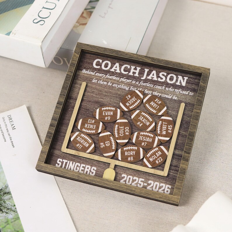 Może przedstawiać: Kwadratowa drewniana ramka typu shadow box z motywem boiska do futbolu amerykańskiego i bramką. Zawiera napis COACH JASON, STINGERS, 2025-2026 oraz cytat. Wewnątrz znajduje się jedenaście małych, drewnianych wycięć w kształcie piłek z grawerem.