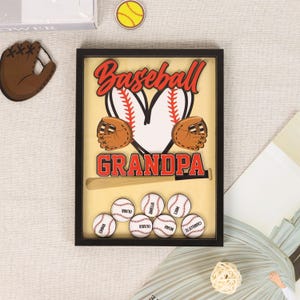Peut inclure: Un souvenir sur le th&egrave;me du baseball encadr&eacute; avec le mot "Baseball" en script rouge au-dessus de "Grandpa". Le design comprend une balle de baseball, des gants et des balles de baseball avec des noms. Le cadre est noir.