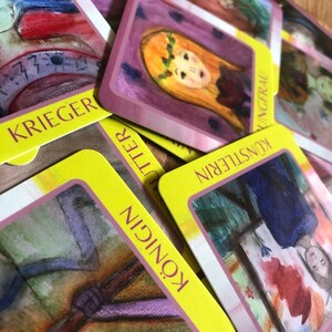Peut inclure: Un ensemble de cartes de tarot avec des illustrations colorées et du texte allemand. Les cartes présentent des images de personnes et d'objets, avec des bordures jaunes et des fonds roses. Les cartes sont disposées sur une surface en bois.
