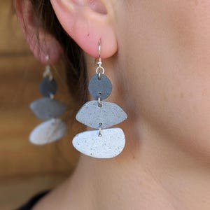 May include: Gray and white stone-shaped dangle earrings. Each earring features three stacked, asymmetrical stone shapes in varying shades of gray and white, suspended from a silver hook. The earrings have a minimalist, modern design.