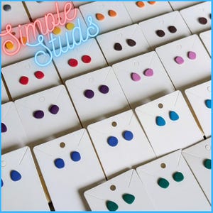 May include: Assortment of colorful polymer clay earrings displayed on white cards. The earrings are in various shapes, including circles, ovals, and triangles. The text "Simple Studs" is displayed in neon pink and blue letters.