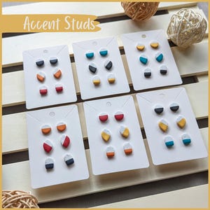 May include: Six cards displaying various sets of colourful geometric accent studs. Each card showcases three pairs of earrings with a white, grey, and coloured design.
