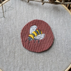 Könnte beinhalten: Handgestickte Stickerei einer Biene auf einem maroonfarbenen runden Patch, der in einem hölzernen Stickrahmen befestigt ist. Die Biene hat gelbe und schwarze Streifen, weiße Flügel und schwarze Details. Der Hintergrund ist ein hellgrauer Stoff.