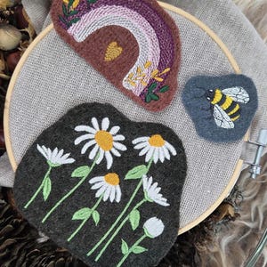 Könnte beinhalten: Gestickte Filzaufnäher mit Regenbogen, Gänseblümchen und einer Biene. Der Regenbogen-Aufnäher hat lila, rosa und weiße Streifen. Der Gänseblümchen-Aufnäher hat weiße Blütenblätter und gelbe Zentren. Der Bienen-Aufnäher ist gelb und schwarz.