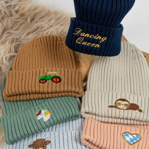 Op de afbeelding: Een verzameling geribde gebreide mutsen in diverse kleuren, waaronder marineblauw, bruin, groen en roze. Sommige mutsen hebben geborduurde ontwerpen zoals een tractor, eenhoorn, luiaard en een Schotse hooglander. Eén marineblauwe muts heeft "Dancing Queen" in goud geborduurd.