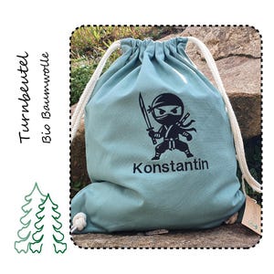 Könnte beinhalten: Eine hellblaue Turnbeutel mit einem schwarzen Ninja-Motiv und dem Namen Konstantin darauf gedruckt. Die Tasche ist aus Bio-Baumwolle.