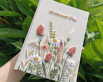 Può includere: Un diario in lino bianco con fiori selvatici ricamati e il testo "Wild and Free" in filo dorato.