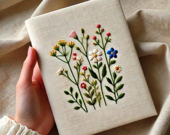 May include: A white linen notebook with a hand-stitched embroidery design of colorful wildflowers. The flowers are in shades of blue, pink, yellow, and green.