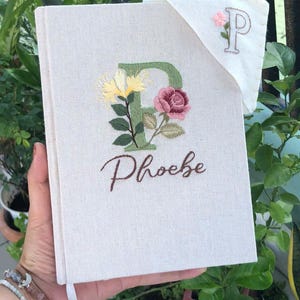 Pode incluir: Diário creme com design floral bordado e o nome "Phoebe". A letra "F" é bordada em verde com flores amarelas e rosas. Um marcador de página a condizer com a letra "P" também é visível.