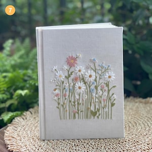 May include: A hardback journal with a light beige linen cover. The cover features an embroidered floral design with white, pink, and blue daisies and other wildflowers. The journal is closed and sits on a woven mat.