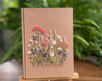 Può includere: Un diario color beige con un ricamo floreale. Il ricamo presenta fiori rossi, rosa, bianchi e blu con steli e foglie verdi. Il diario è esposto su un piccolo cavalletto di legno.