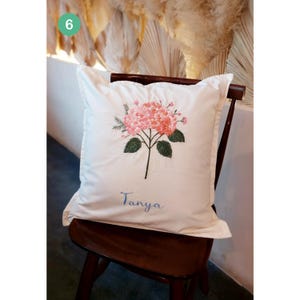 May include: White cushion with a pink flower embroidered on it. The cushion has the name "Tanya" embroidered on it in blue thread.