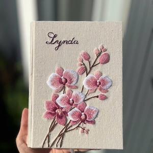 May include: A beige journal with an embroidered floral design. The cover features pink and white orchids with brown stems and the name "Lynda" embroidered in cursive. A white ribbon bookmark is visible.