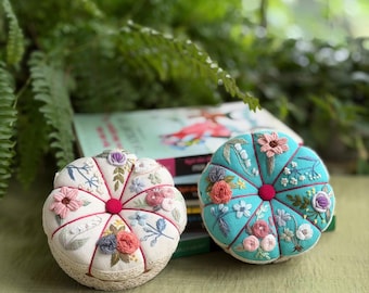 May include: Two round, tufted pincushions featuring intricate floral embroidery. One is cream-colored with pink, blue, and purple thread details, while the other is bright turquoise with similar floral motifs. Both have a central button.