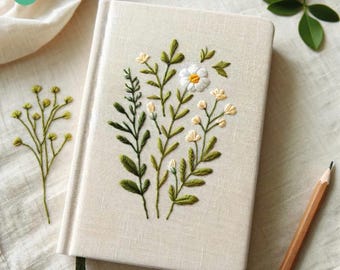 May include: A white linen notebook with green embroidery of flowers and leaves. The notebook has a green ribbon bookmark.