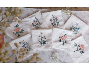 May include: A collection of white linen handkerchiefs, each embroidered with a different floral design. The embroidery features various colors, including pink, purple, blue, and green, depicting roses, lavender, daisies, and other flowers. The handkerchiefs are arranged on a floral patterned surface.