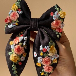May include: A black hair bow featuring embroidered flowers in shades of pink, yellow, red, and blue, with green leaves. The bow is made of a textured fabric. The floral embroidery is detailed.