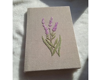 May include: A beige canvas with a hand-stitched lavender flower design in shades of purple and green.